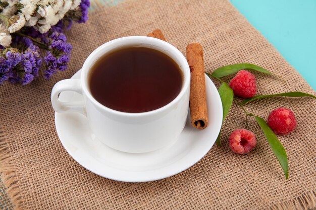 Close up view of cup of tea and cinnamon on saucer with raspberries and leaves and flowers on sackcloth