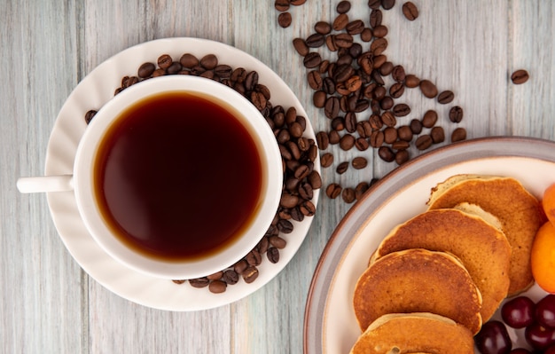 Close-up view of cup of tea and coffee beans on saucer with plate of pancakes cherries apricots on wooden background