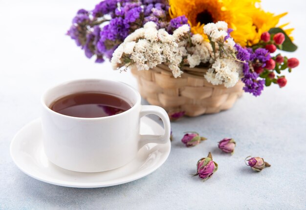 Close up view of cup of tea on saucer and flowers in basket and on white surface