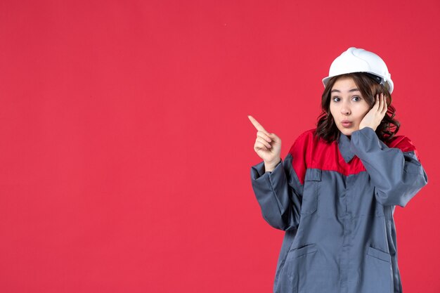 Close up view of curious female builder in uniform with hard hat and pointing something on the right side on isolated red wall