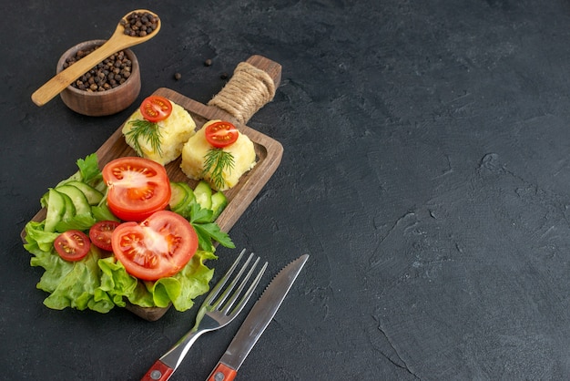 Close up view of cut fresh tomatoes and cucumbers cheese on wooden board cutlery set on dark color towel on the right side on black surface