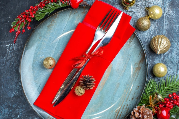 Vista ravvicinata del set di posate con nastro rosso su un tovagliolo decorativo su un piatto blu e accessori natalizi su sfondo scuro