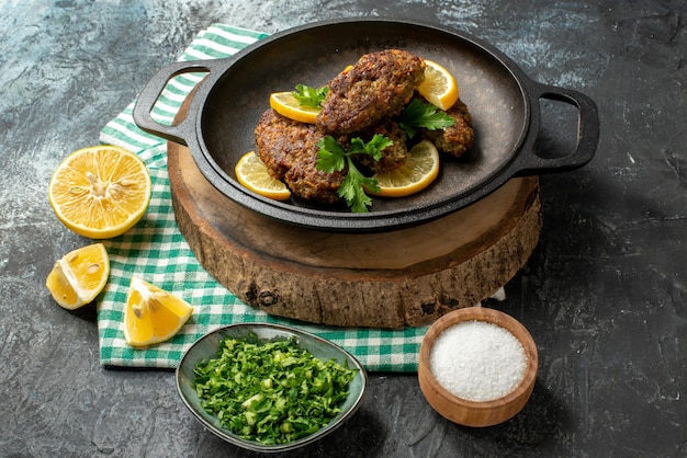 Close up view of delicious cutlets served with greens and lemon in black pan on wooden board salt on green stripped towel on dark color background