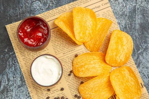 Close up view of delicious homemade chips and pepper bowl mayonnaise ketchup on newspaper on gray table