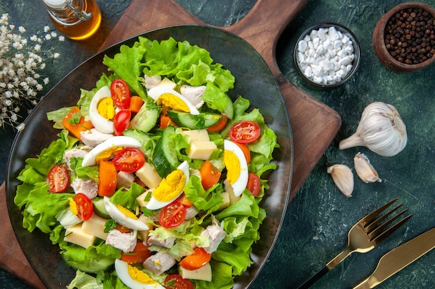 Close up view of delicious salad with many fresh ingredients on wooden cutting board spices oil bottle garlics cutlery set on black green mix colors background
