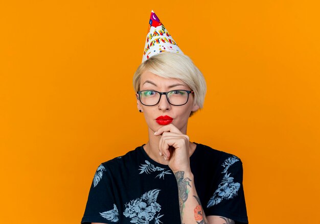 Close-up view of doubtful young blonde party girl wearing glasses and birthday cap looking at camera touching chin isolated on orange background with copy space