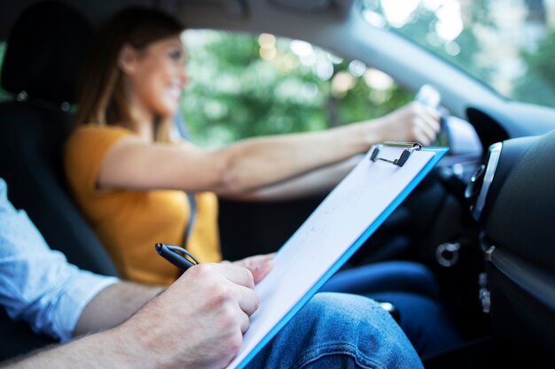 Close up view of driving instructor holding checklist while in background female student steering and driving car