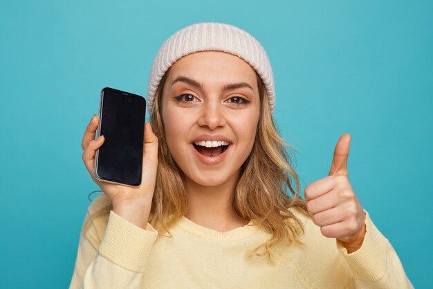 Крупным планом возбужденная молодая девушка в зимней шапке, держащая мобильный телефон, показывает палец вверх