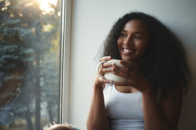 Close up view of fashionable cute young African American female in white tank top having rest indoors, holding large cup of hot tea, smiling broadly, daydreaming, spending nice time at home alone