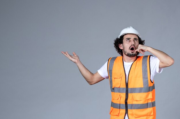 Close up view of focused male architect in warning vest with safety helmet and pointing something on the right side making call me gesture on gray wave wall