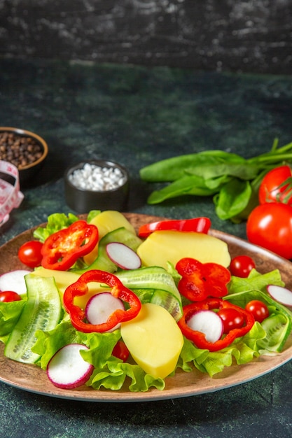 Close up view of fresh peeled cut potatoes with red pepper radishes green tomatoes in a brown plate and meters spices on green black mix colors surface