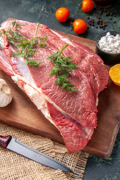 Close up view of fresh raw red meats green garlics lemon salt on brown wooden cutting board knife on nude color towel tomatoes pepper on dark color background