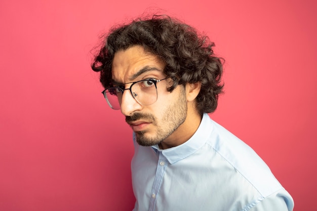 Close-up view of frowning young handsome man wearing glasses standing in profile view looking at front isolated on pink wall