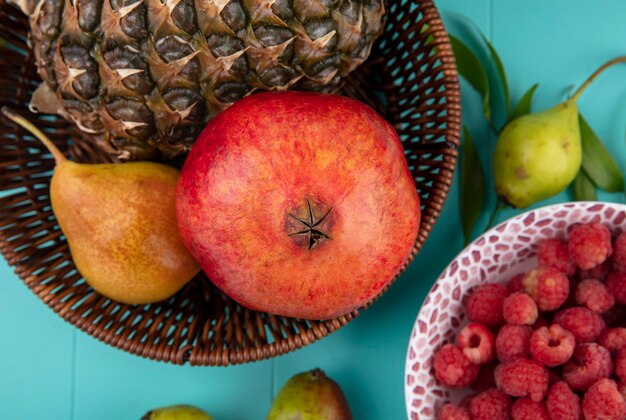 Close up view of fruits as pineapple pomegranate peach in basket and bowl of raspberry with peaches and leaves on blue surface