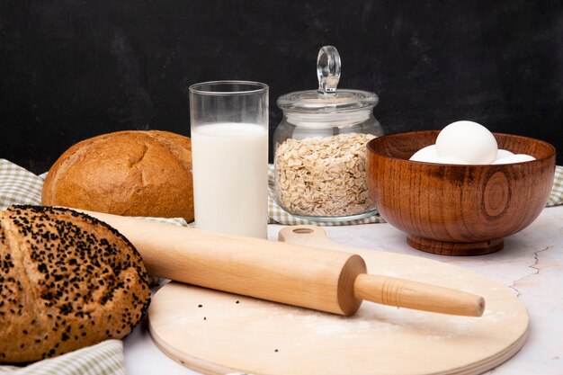 Close-up view of glass of milk and bowl of eggs with breads oat-flakes rolling pin on cutting board on white surface and black background