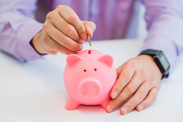 Close up view of the hand of a man placing a coin into the slot of a piggy bank in a savings and investment concept