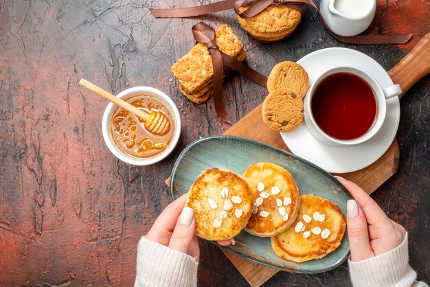 Close up view of hand taking tray with fresh pancakes a cup of black tea on a wooden cutting board honey stacked cookies milk on a dark surface