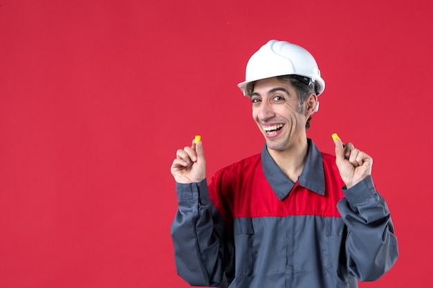 Close up view of happy young builder in uniform with hard hat and holding earplugs on isolated red wall