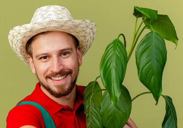 Close-up view of happy young handsome slavic gardener in uniform and hat holding plant  isolated on olive green wall