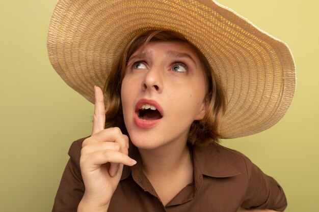 올리브 녹색 벽에 격리된 채 해변 모자를 쓰고 가리키는 인상을 받은 젊은 금발 소녀의 클로즈업 보기