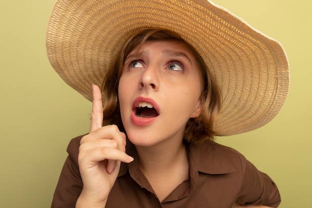 Close-up view of impressed young blonde girl wearing beach hat looking and pointing up isolated on olive green wall
