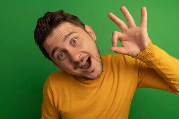 Close-up view of impressed young blonde handsome man looking  doing ok sign isolated on green wall