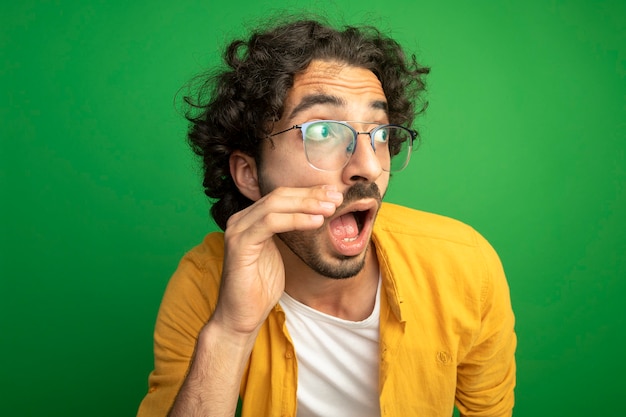 Foto gratuita vista ravvicinata di impressionato giovane uomo bello con gli occhiali guardando il lato toccando il viso isolato sulla parete verde