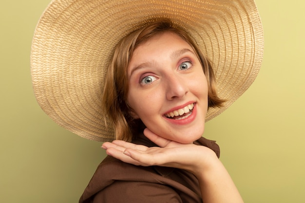 Close-up view of joyful young blonde girl wearing beach hat standing in profile view looking  touching chin isolated on olive green wall