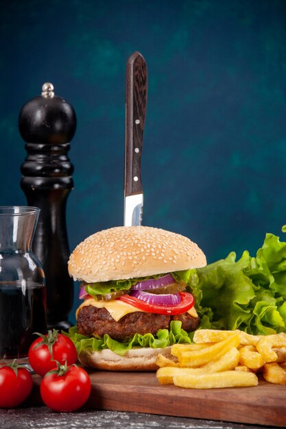 Free photo close up view of knife in meat sandwich and fries tomatoes with stem green bundle on wooden board sauce ketchup on dark blue surface