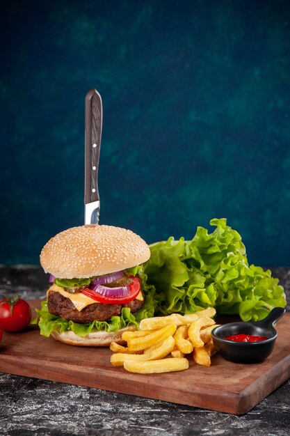 Close up view of knife in meat sandwich and fries tomatoes with stem pepper on wooden board ketchup on dark blue surface