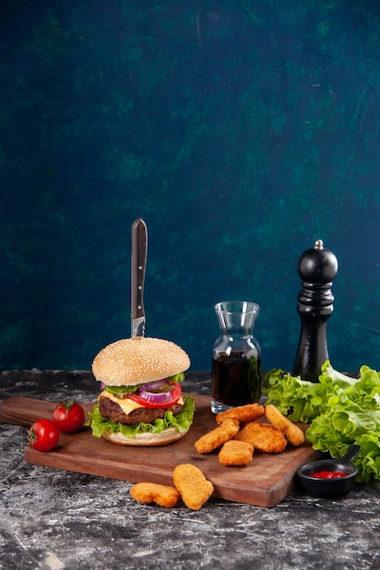 暗い青色の表面にソース ケチャップを木の板に茎唐辛子とおいしい肉のサンドイッチとチキン ナゲット トマトのナイフのクローズ アップ ビュー