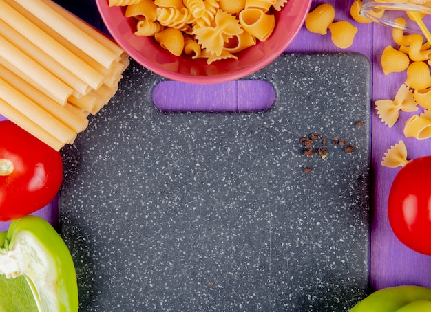 Close-up view of macaronis as bucatini rotini and others with pepper tomato around cutting board on purple surface