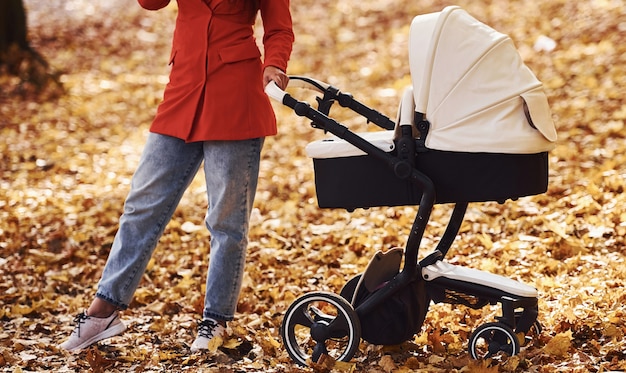 Крупным планом вид. мама в красном пальто гуляет с ребенком в детской коляске в парке осенью.