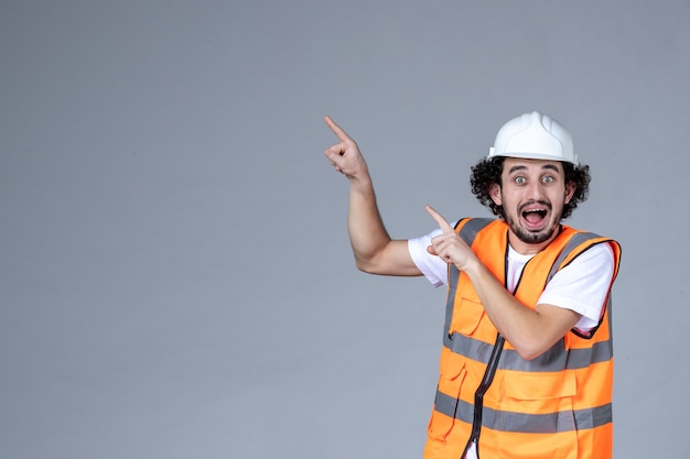 Close up view ofnervous emotional male architect in warning vest with safety helmet and pointing something on the right side on gray wave wall