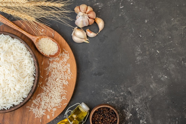 Vista ravvicinata di riso lungo perfetto in pentola marrone e su tavola di legno cucchiaio di aglio cucchiaio di pepe caduto bottiglia di olio su sfondo grigio