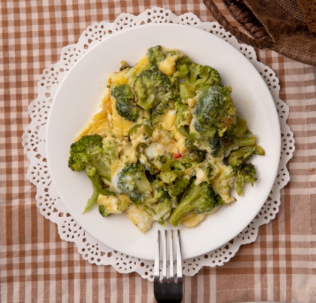 Close-up view of plate of meal with eggs and broccoli and fork on paper doily on plaid cloth background