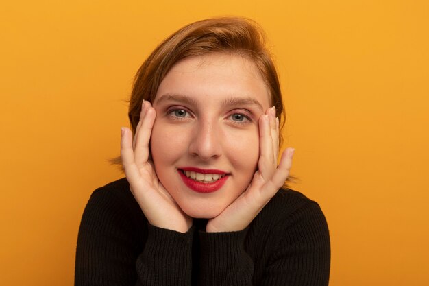 Close-up view of pleased young blonde girl putting hands on face  isolated on orange wall