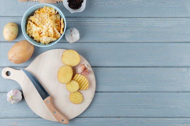 Close up view of potato slices and garlic with knife on cutting board and crisps on wooden background with copy space
