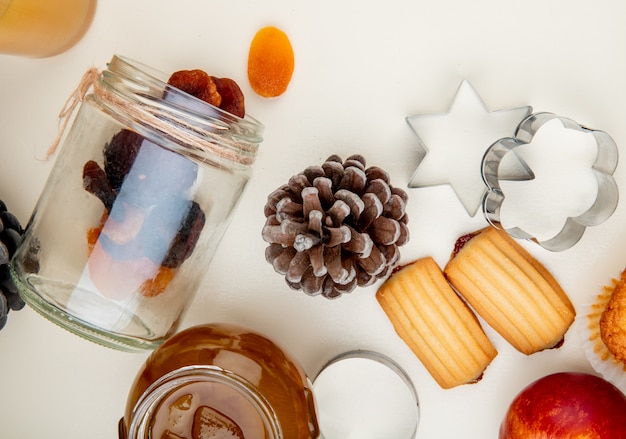 Close-up view of raisins spilling out of jar and pinecone with peach jam and cookies on white table