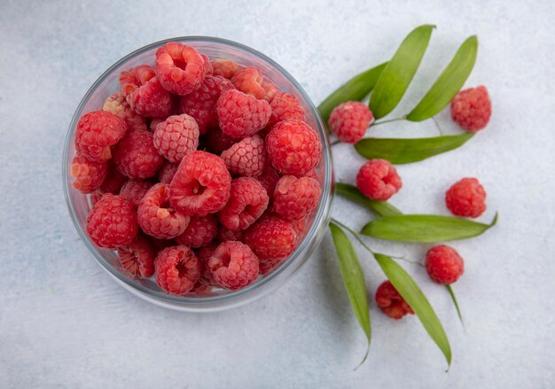 Close up view of raspberries in glass bowl with leaves and raspberries on white surface