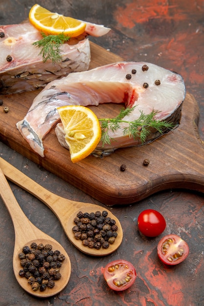 Close up view of raw fishes and pepper on wooden cutting board lemon slices tomatoes on mix color surface