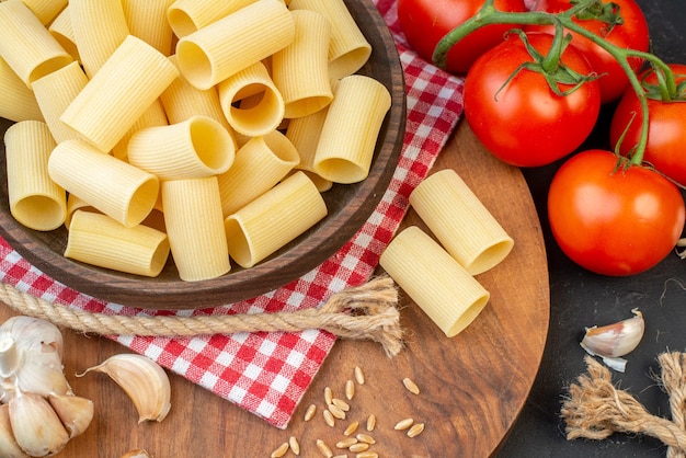 Close up view of raw pastas inside and outside a brown bowl on red stripped towel garlics rice on round wooden board tomatoes rope on black background