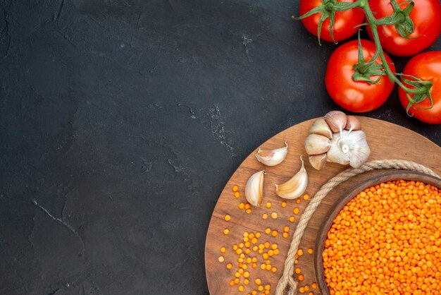 Close up view of red lentil in a brown bowl rope garlics on wooden round board and tomatoes with stems on the left side on black background