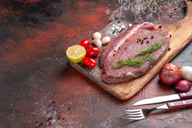 Close up view of red meat on wooden cutting board and garlic green lemon onion fork and knife on dark background