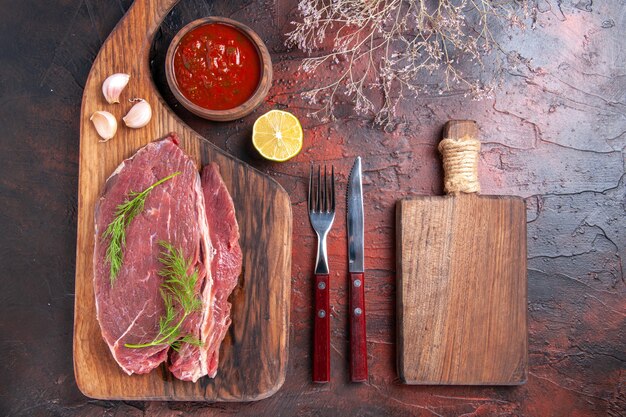 Close up view of red meat on wooden cutting board and ketchup in small bowl fork and knife on dark background