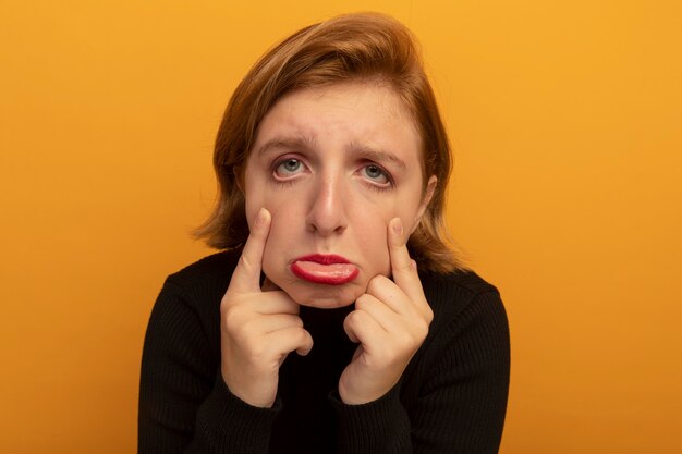Close-up view of sad young blonde woman looking at front pulling down eye lids isolated on orange wall