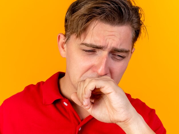 Close-up view of sad young handsome blonde ill man looking down touching mouth isolated on orange wall