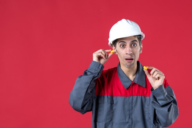 Крупным планом вид шокированного молодого строителя в униформе с каской и держащего беруши на изолированной красной стене