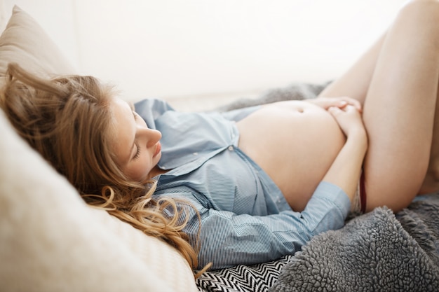 Крупным планом вид спящей молодой светловолосый беременной женщины в голубой рубашке, лежа в удобной кровати, держа ее живот руками во время сна. Драгоценные моменты родительства.