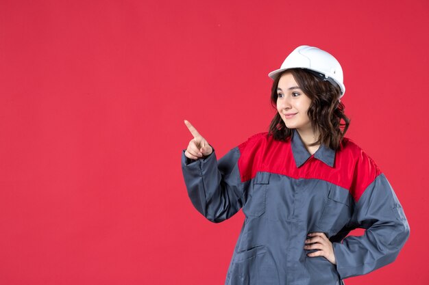 Close up view of smiling female builder in uniform with hard hat and pointing something on isolated red wall